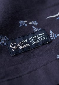 Tela azul marino con un patrón floral en azul claro, con una etiqueta negra que dice "Superdry Clothing Co" y sus instrucciones de cuidado, cosida a ella.