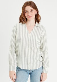 Chemise rayée à manches longues en vert clair et blanc, fabriquée en tissu léger. Elle possède une poche et un col. Détails de boutons sur les manches.