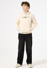 Felpa beige con cappuccio e tasca frontale, con "ALTERNATIVE LEGENDS" stampato in nero; abbinata a jeans neri e sneakers bianche.