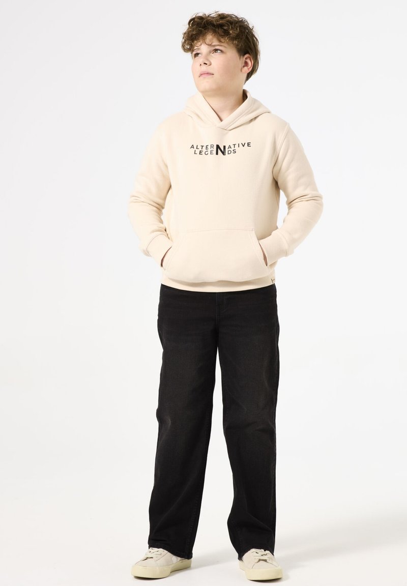 Felpa beige con cappuccio e tasca frontale, con "ALTERNATIVE LEGENDS" stampato in nero; abbinata a jeans neri e sneakers bianche.