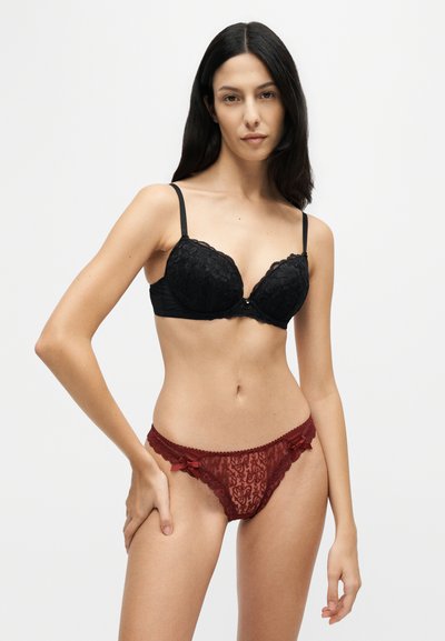 Femme aux longs cheveux foncés portant un soutien-gorge en dentelle noire et une culotte en dentelle rouge, debout sur un fond clair uni.