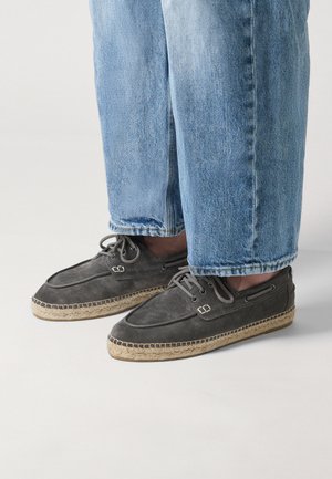 Person trägt graue Espadrilles aus Wildleder mit Schnürsenkeln und hellblauen Jeans, steht auf einem weißen Boden.
