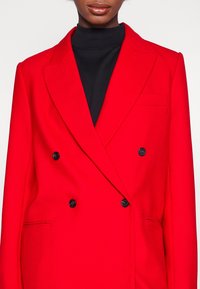 Blazer rojo de doble botonadura hecho de una tela texturizada, con solapas de muesca, dos botones negros y un bolsillo en el pecho izquierdo.