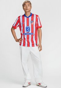 Tricou de fotbal cu dungi roșii și albastre, cu mâneci scurte, având un guler albastru și un logo. Purtați cu pantaloni albi și adidași.