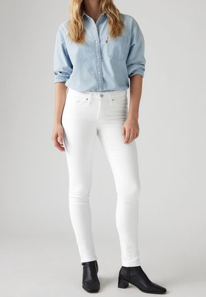 Femme portant une chemise bleu clair boutonnée, un jean skinny blanc et des bottines à talons noires, debout devant un fond gris uni.