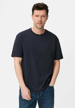 Man in een effen zwart T-shirt met korte mouwen en blauwe spijkerbroek, staand met één hand in zijn zak tegen een witte achtergrond.