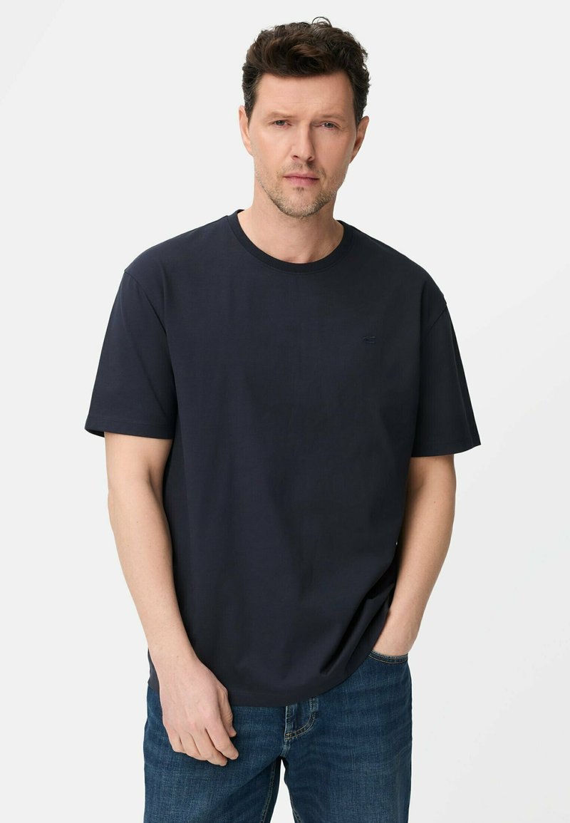 Man in een effen zwart T-shirt met korte mouwen en blauwe spijkerbroek, staand met één hand in zijn zak tegen een witte achtergrond.