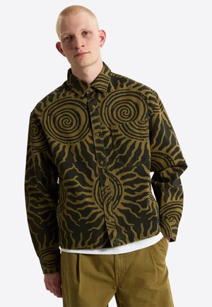 Junger Mann mit heller Haut und kurzen blonden Haaren, der ein grün-schwarz spiralförmiges Hemd mit Knöpfen und hellbraune Hose trägt.