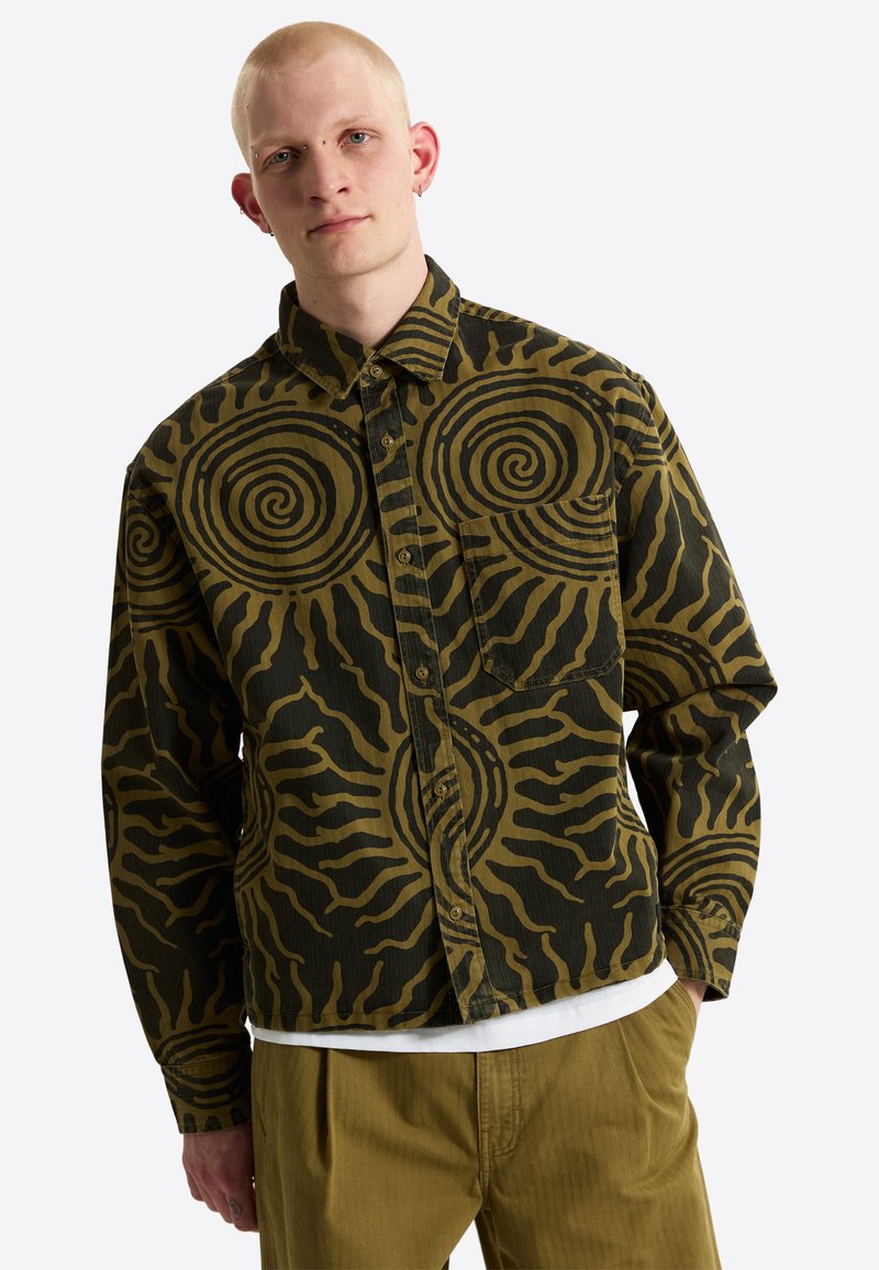 Young man with light skin and short blonde hair wearing a green and black spiral-patterned button-up shirt and light brown pants.