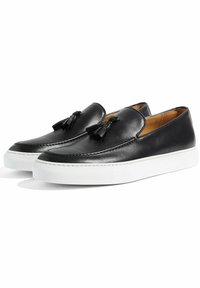 Scarpe slip-on in pelle nera con dettagli a nappina, dotate di suola in gomma bianca e cuciture lungo la parte superiore. Texture liscia.