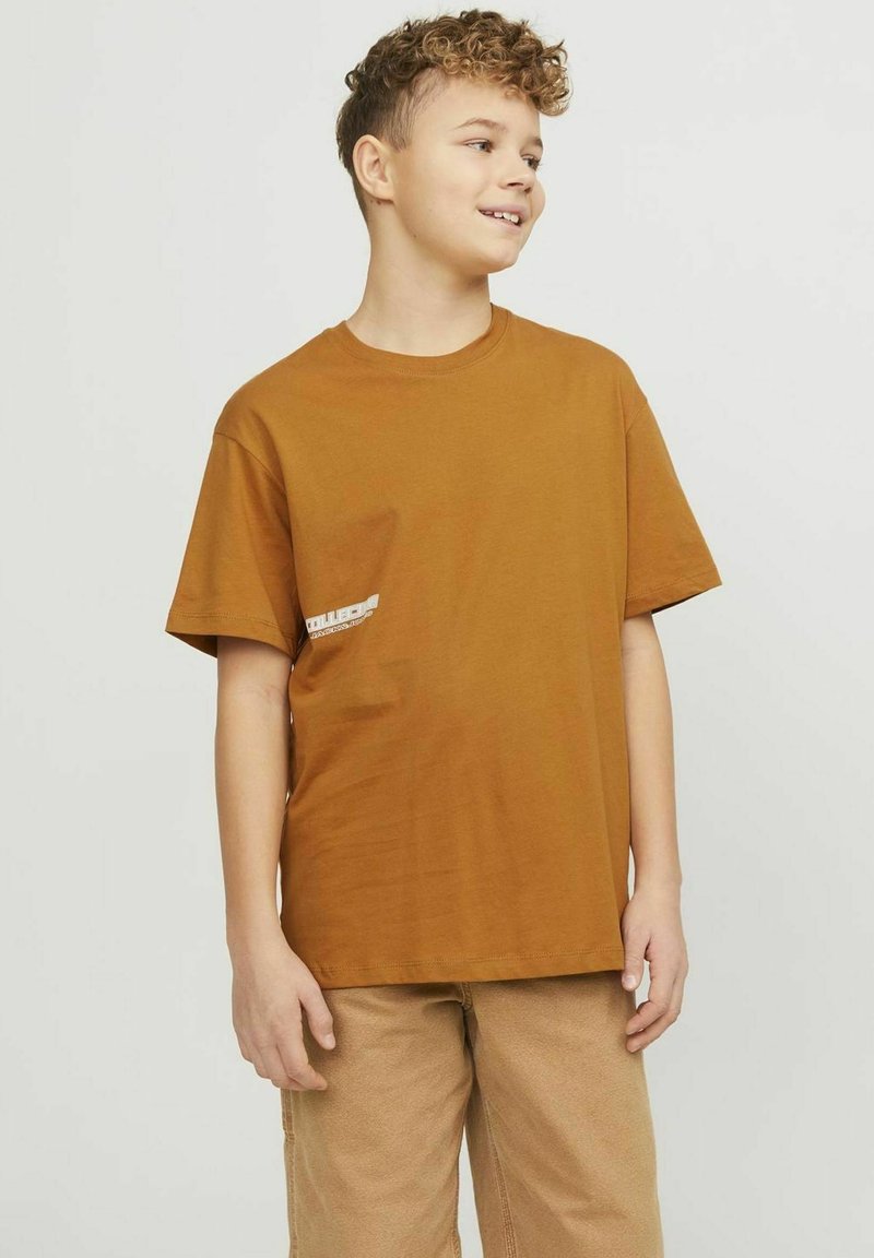 Brown cotton t-shirt with a relaxed fit, short sleeves, and a small white logo on the left chest. Paired with beige pants.