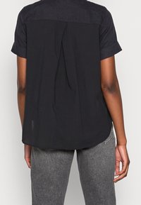 Blusa nera a maniche corte con orlo arrotondato, caratterizzata da una piega sul retro e un tessuto a contrasto sulla spalla.