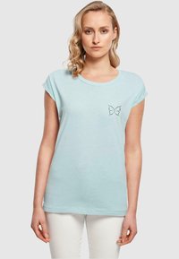 Lys blå kortermet t-skjorte med rund hals og en liten brodert sommerfugl-design på venstre brystområde.