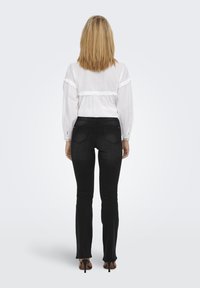 Hvit bluse med volanger på ermene og detaljer på ryggen, kombinert med svarte tettsittende jeans. Modell iført svarte stilettohæler.