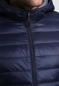 Marinblå puffjacka med quiltat mönster, har en framsidan dragkedja, hög krage och slät textur. Lätt material som är lämpligt för lager-på-lager.