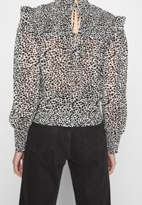 Genomskinlig blus med ett svart och brunt prickigt mönster, med en volangkrage, smockad midja och långa puffärmar.