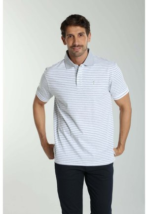 Homme aux cheveux courts et foncés portant un polo rayé gris clair et blanc et un pantalon noir, debout devant un fond uni.