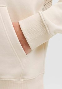 Manche de sweatshirt couleur crème montrant un poignet côtelé, une texture de tissu lisse et une poche. Détails de couture subtils et design minimaliste.