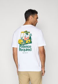 White short-sleeve t-shirt featuring a colorful graphic of a man in a taxi with the text "Patience Required" in green. Casual fit.