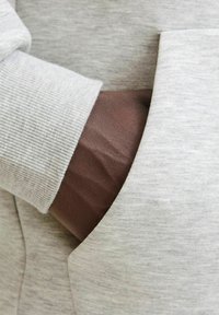 Helles graues Sweatshirt mit gerippten Bündchen. Eine dunkelbraune Hand befindet sich in der Kängurutasche, die die Textur des Stoffes und die Nähdetails zur Schau stellt.