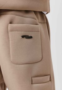Beige joggingbroek van zachte stof, met een grote zak op de rechterdij en een zwart metalen logo-accent.
