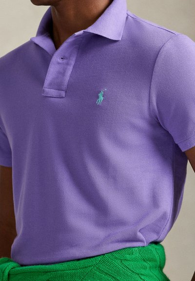Mann trägt ein violettes Poloshirt mit türkisfarbenem Logo und ein grünes gestricktes Kleidungsstück um die Taille gebunden vor neutralem Hintergrund.