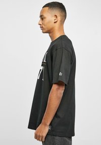 Tricou negru din bumbac, cu mâneci scurte și decolteu rotund, având text alb pe față și un mic logo cu stea pe mâneca stângă.