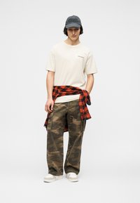 Νεαρός άνδρας που φοράει γκρι καπέλο, κρεμ t-shirt McLaren, κόκκινο και μαύρο καρό πουκάμισο δεμένο γύρω από τη μέση, παντελόνι καμουφλάζ cargo και λευκά αθλητικά παπούτσια.