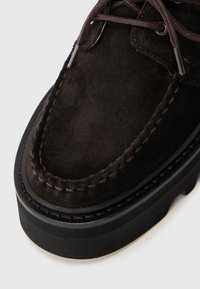 Scarpa in suede marrone scuro con punta arrotondata, cuciture decorative e una spessa suola in gomma nera. Presenta lacci marroni e occhielli in argento.