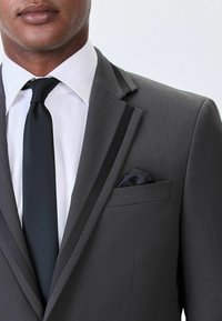 Chaqueta de traje gris texturizada con solapa de satén negro, camisa blanca y una corbata negra. El pañuelo de bolsillo presenta un patrón oscuro, realzando el diseño general.
