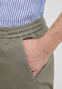 Pantalones cortos de algodón verde olivo claro con una cinturilla elástica, bolsillo lateral y una textura suave, combinados con una camisa a rayas azul y blanca.