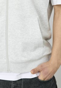 Haut de hoodie zippé gris clair en tissu doux avec poches kangourou, ourlet côtelé, et t-shirt blanc superposé en dessous.