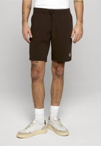 Bruine katoenen shorts met een elastische tailleband en trekkoord. Heeft een logodetail op het onderbeen. Draagt witte sneakers en sokken.