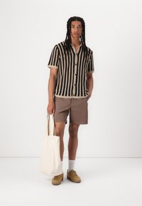 Maglietta a maniche corte lavorata a maglia con strisce verticali nere e beige, abbinata a pantaloni corti marroni, sneakers beige e una borsa a tracolla crema.