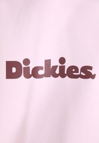 Sfondo rosa con il grande e audace testo "Dickies" in marrone scuro. Il tessuto appare liscio con una leggera lucentezza, senza ulteriori motivi o accenti.