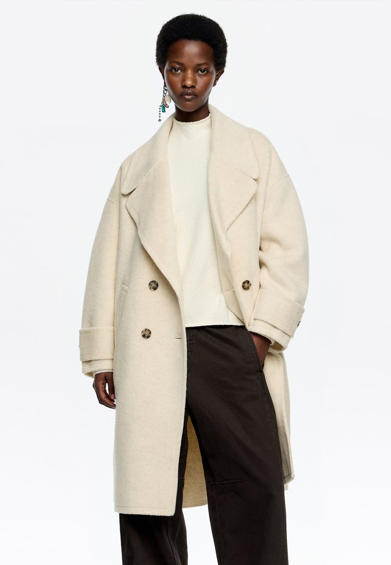 Cappotto doppiopetto in misto lana beige con colletto a risvolto, grandi bottoni e tasche laterali, indossato sopra un top crema e pantaloni scuri.