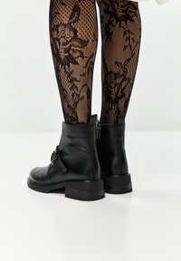 Bottines en cuir noir avec un bout arrondi, une boucle décorative et un petit talon carré, associées à des collants noirs à motif en dentelle.