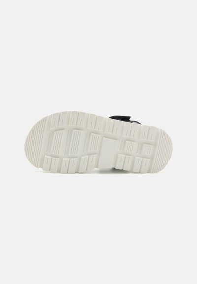 Zapatilla negra con una gruesa suela de goma blanca. La suela exterior presenta un patrón de huella horizontal y vertical para una mejor tracción.