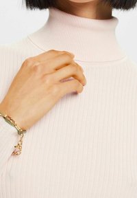 Top de cuello alto acanalado de color rosa claro con un diseño ajustado. La mano está posicionada cerca del escote, mostrando una textura suave y un brillo sutil.
