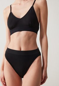 Sutien negru și slip de bikini cu talie înaltă, realizat dintr-un material neted. Sutienul are un decolteu în V și detalii plisate; talia este ribbită.