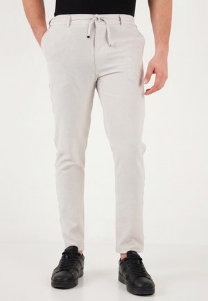 Hombre con pantalones ajustados de color beige claro con cintura de cordón y zapatillas negras, de pie frente a un fondo liso.
