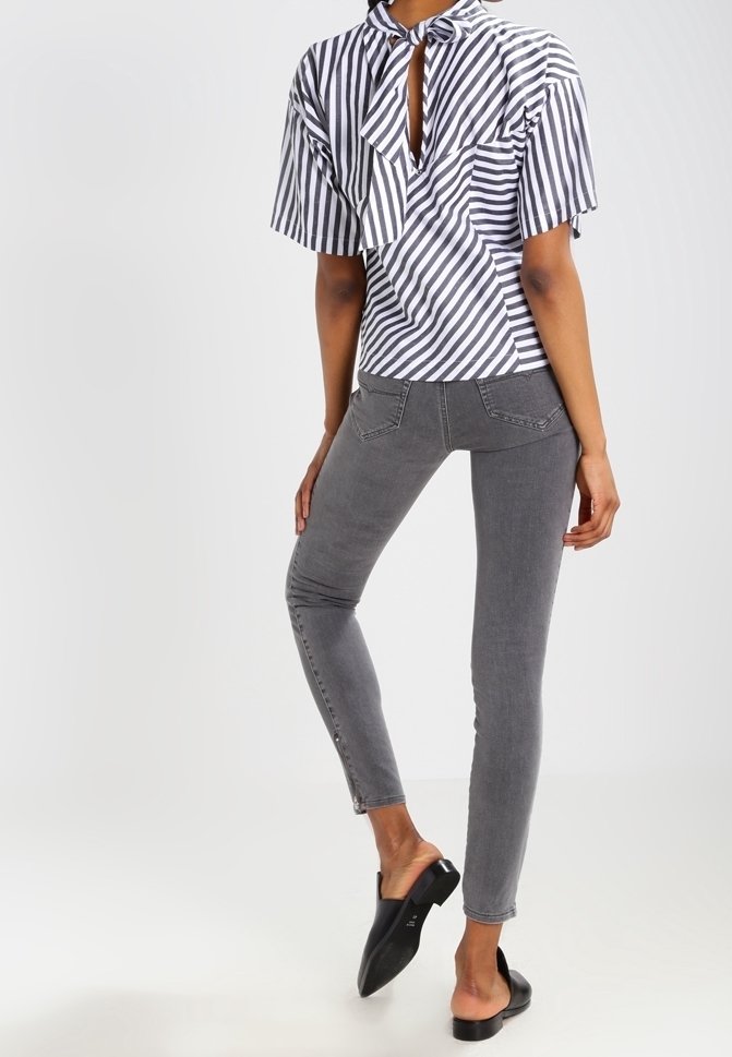 Femme portant un chemisier rayé noir et blanc à manches courtes, un jean skinny gris et des talons noirs enfilés, debout avec une jambe légèrement levée.