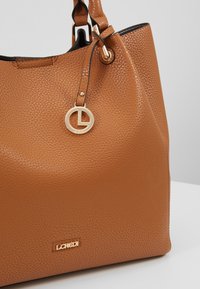 Sac à main en cuir marron texturé avec un charm circulaire doré et une plaque logo portant l'inscription L.CREDI sur le devant.