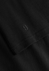 Tela negra con costuras detalladas y un pequeño logo minimalista bordado cerca del dobladillo.