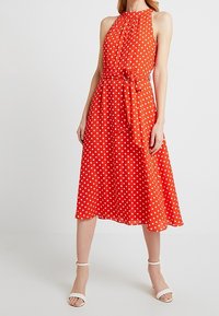 Röd midiklänning med prickar, halterneck, figursydd bodice och knytdetalj i midjan. Flödande tyg med en jämn textur.