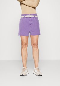 Shorts di denim viola con orlo grezzo, dotati di cintura intrecciata bianca. Indossati con scarpe sportive beige, su sfondo bianco semplice.