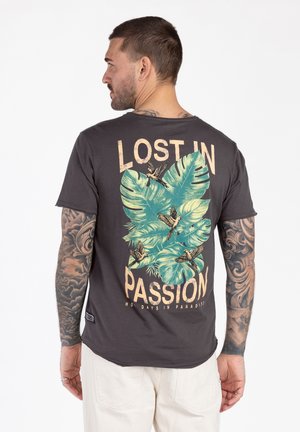 Hombre con tatuajes que lleva una camiseta oscura con grandes hojas verdes, pájaros volando y el texto "Perdido en la pasión, vacaciones en el paraíso" en la espalda.