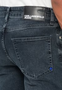 Juodų džinsų šoninis vaizdas su juodu odiniu etiketė, ant kurios užrašyta "KARL LAGERFELD JEANS" ir mažu mėlynu siuvinėtu "K" ant kišenės.