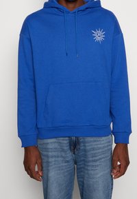 Sweat à capuche bleu en tissu doux, avec une poche avant, une capuche à cordon, et un logo soleil sur la poitrine gauche. Coupe décontractée.