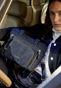 Denim crossbody bag in dark blue with contrasting stitching; features multiple zippers, adjustable strap, and brand logo displayed prominently.